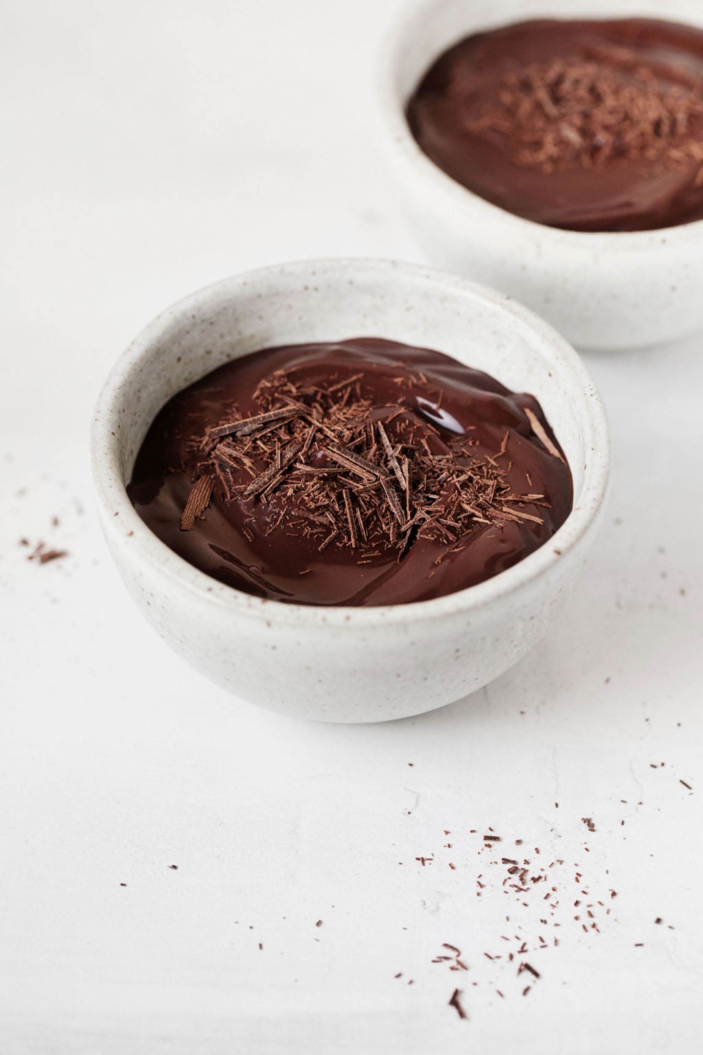 Two small, gray ceramic bowls are filled with a rich, deep mocha colored cashew chocolate pudding and chocolate shavings.