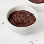 Two small, gray ceramic bowls are filled with a rich, deep mocha colored cashew chocolate pudding and chocolate shavings.