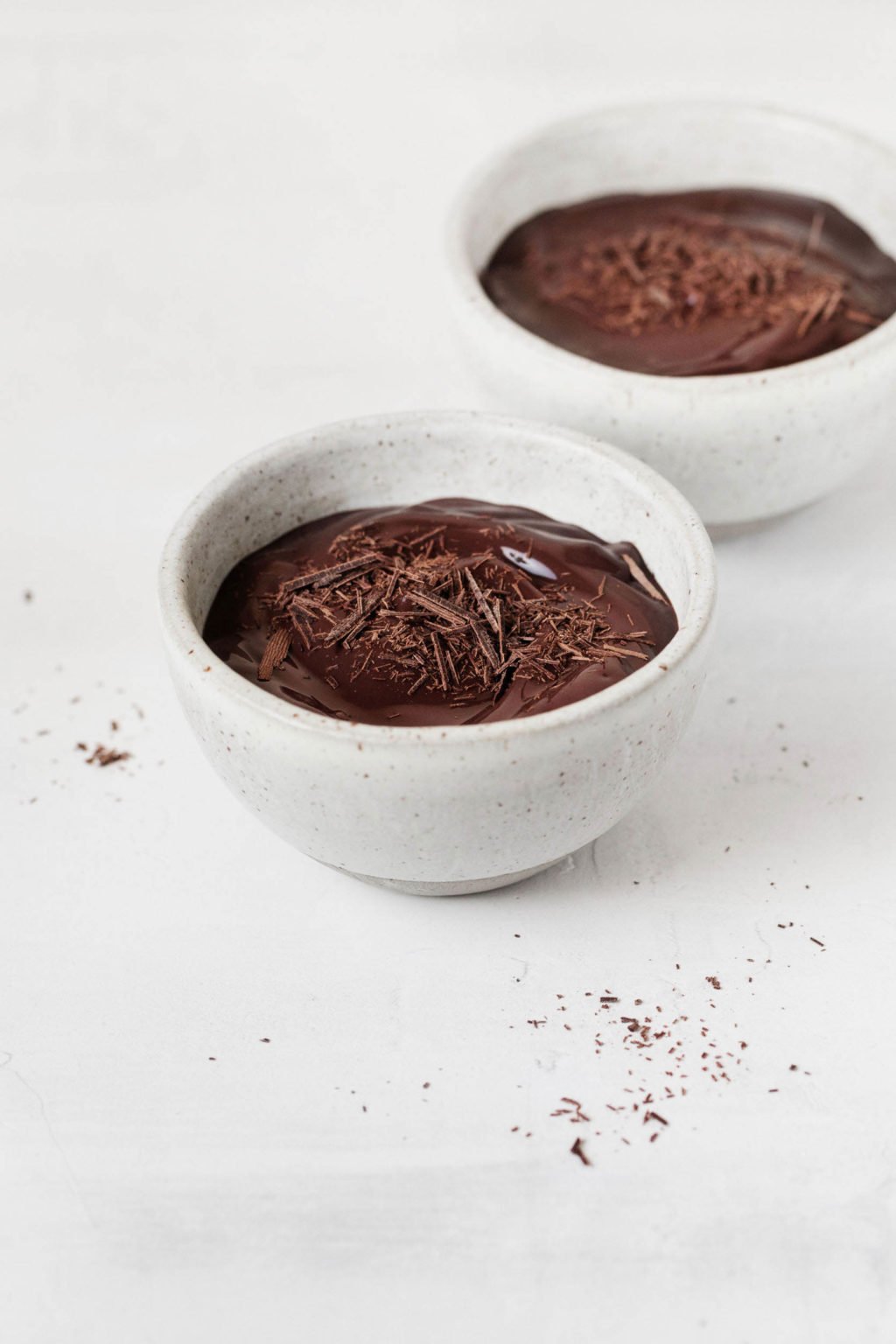 Two small, white ceramic bowls are filled with a vegan cashew chocolate pudding and shaved dark chocolate.