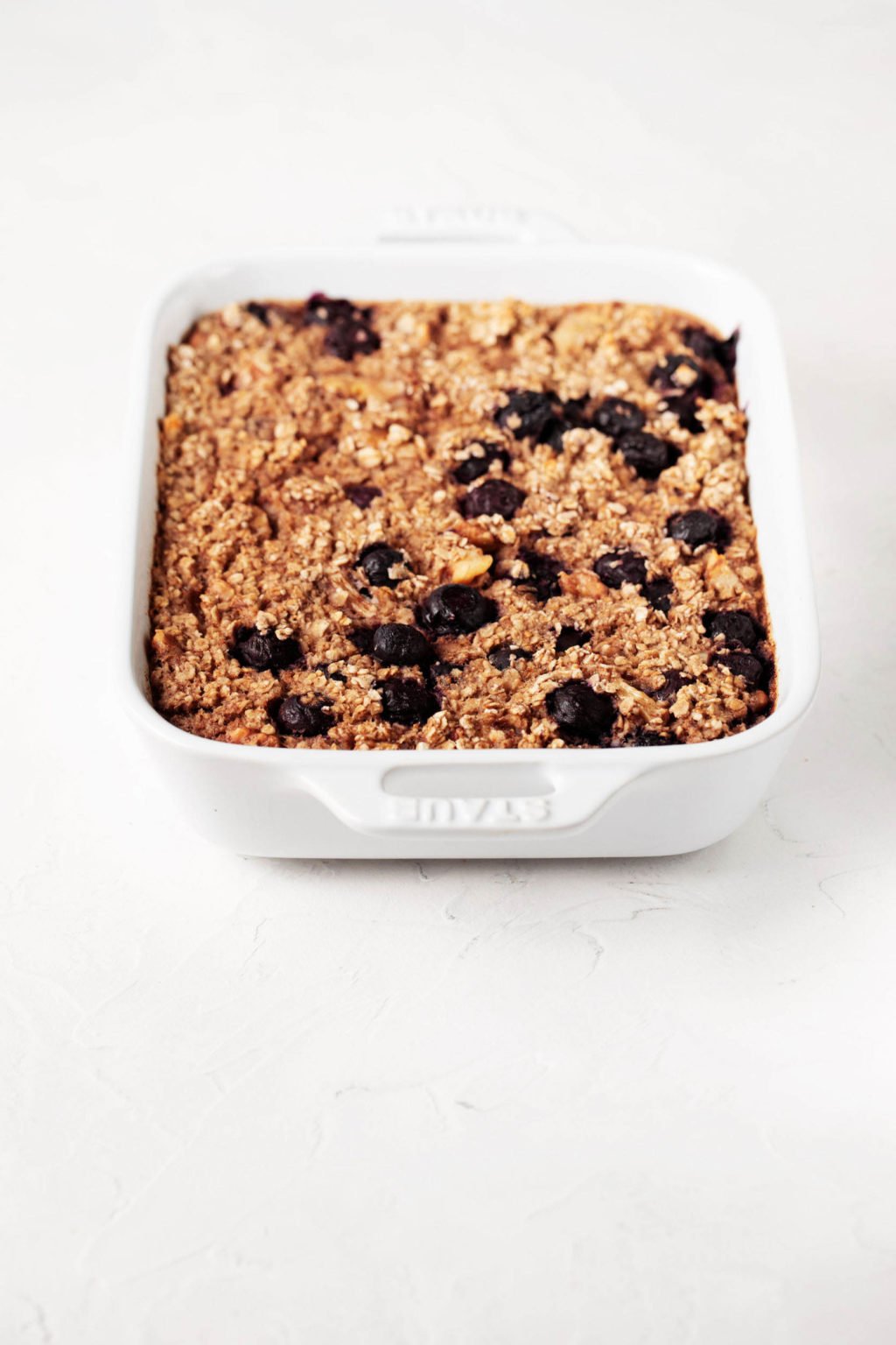 A white, rectangular porcelain dish with handles rests on a white surface. It's filled with a freshly baked, whole grain breakfast dish.