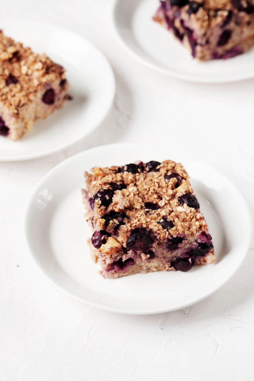 Three small white plates are holding a vegan oat bake with blueberries and banana.