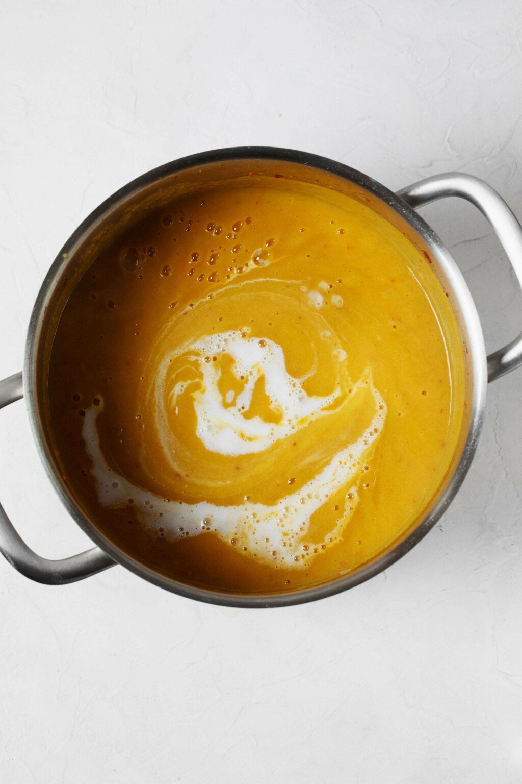 An overhead image of a silver pot, filled with a creamy, orange-hued soup. There's a ribbon of cashew cream running through the soup. 
