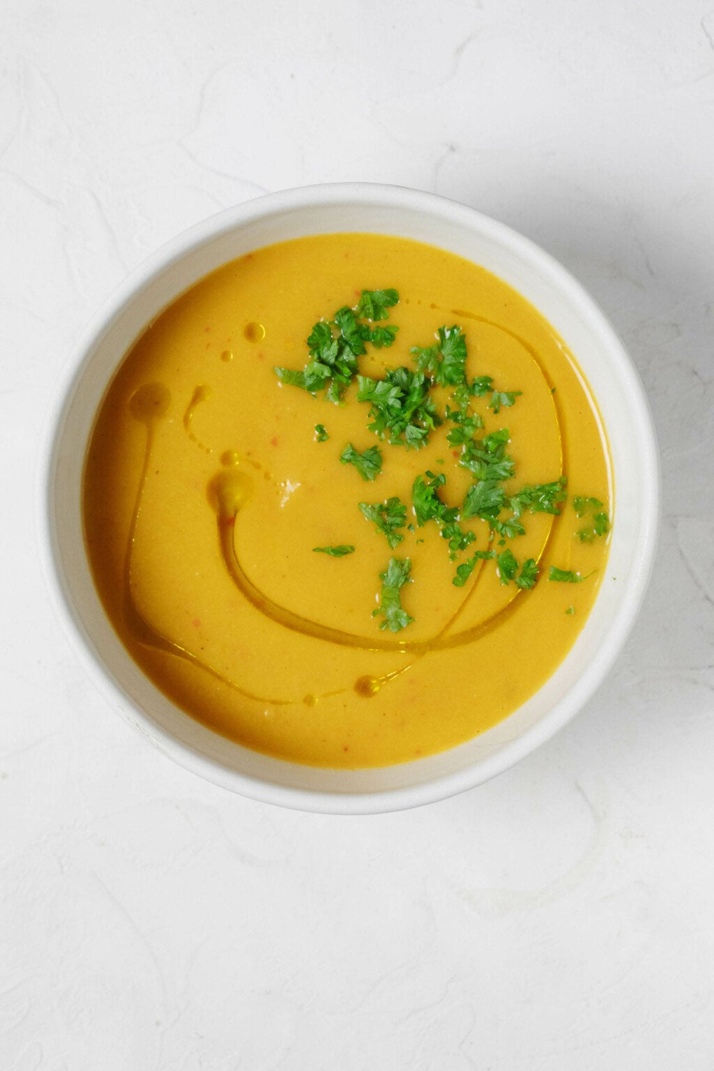 An overhead image of a white bowl, which is filled with an orange-hued creamy curried cauliflower soup, garnished with snipped herbs.