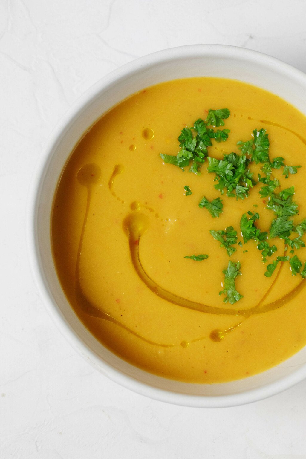 An overhead image of a white bowl, which is filled with an orange-hued creamy curried cauliflower soup, garnished with snipped herbs.