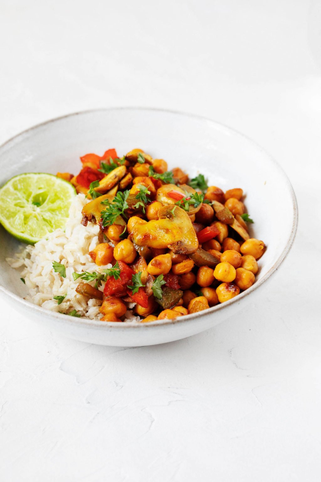 An angled photograph of a white, ceramic bowl with a gray rim. The bowl rests on a white surface and holds a combination of cooked rice and a vegan chickpea scramble.