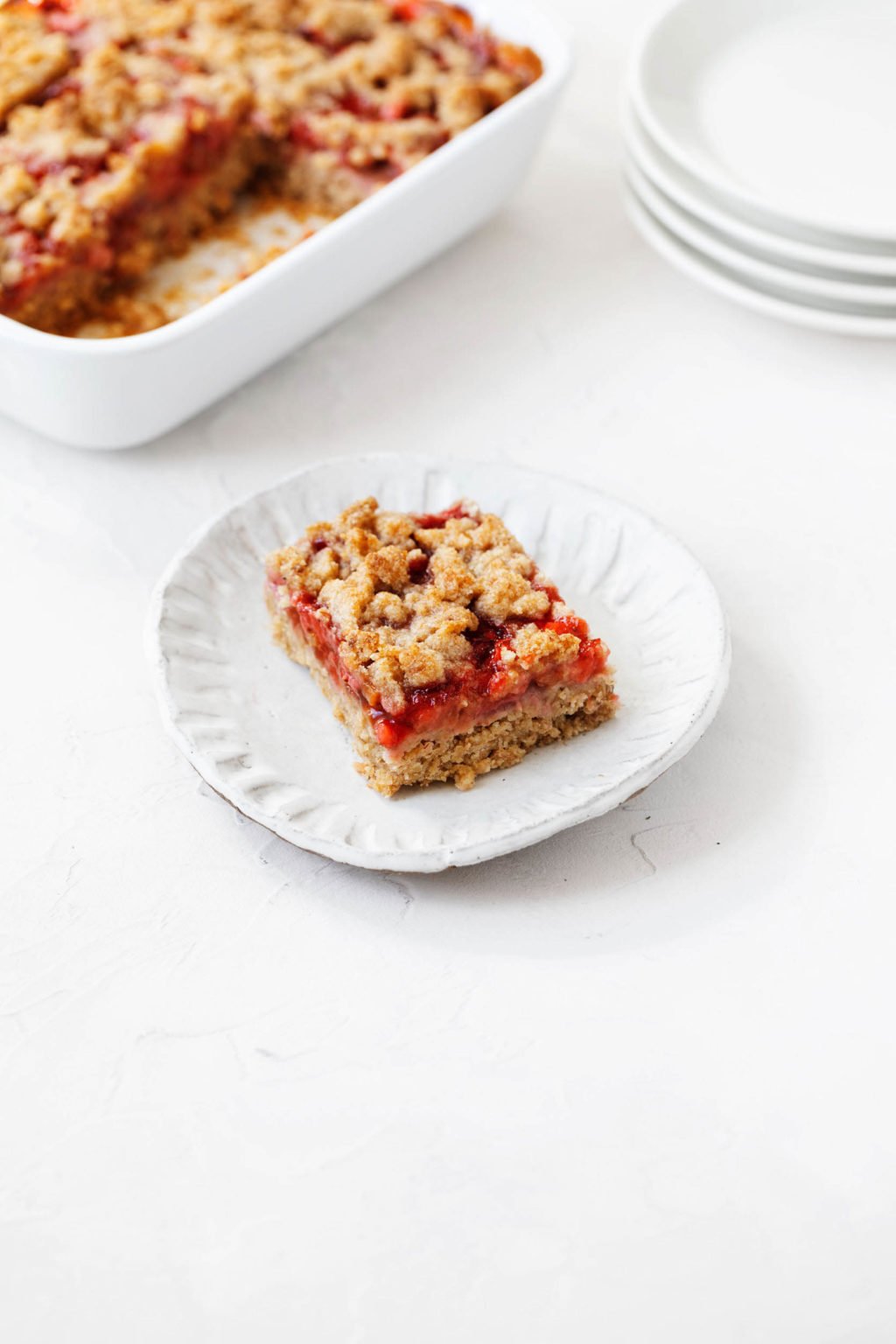 An angled photo of a vegan strawberry rhubarb crumble bar, which is served on a small white dessert plate. There's a rectangular baking dish in the background.
