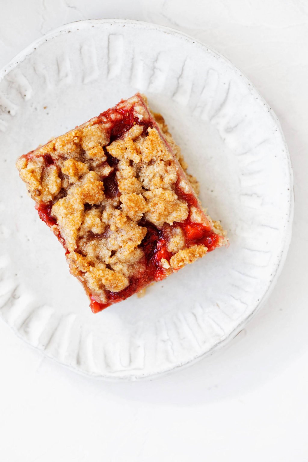 An overhead image of a plant-based strawberry rhubarb crumble bar, resting on a fluted white plate.