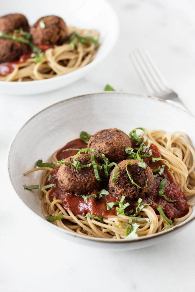A white bowl has been filled with a mix of spaghetti & white bean balls, along with marinara sauce. It's topped with fresh, sliced basil.