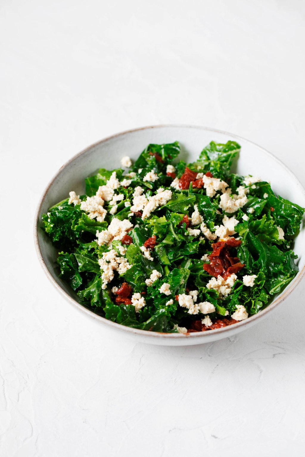 A white, ceramic bowl holds a tofu feta kale salad, which is prepared with small pieces of red sun-dried tomatoes.