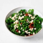 A white, ceramic bowl holds a tofu feta kale salad, which is prepared with small pieces of red sun-dried tomatoes.