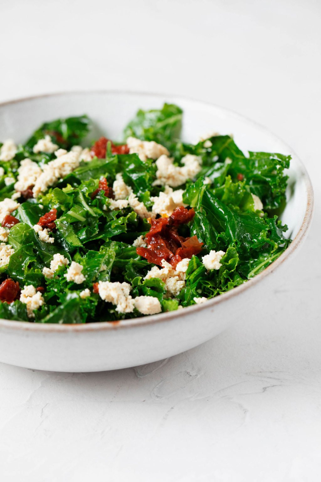 An angled image of a plant-based meal of leafy greens, tomatoes, and a vegan cheese. It's resting on a white surface.