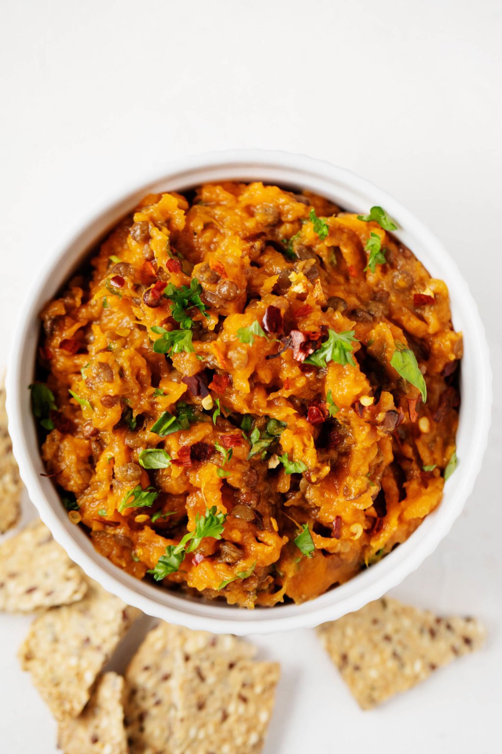 An overhead image of a white bowl of lentil sweet potato salad, which has been sprinkled with chopped parsley. Whole grain crackers lie nearby.