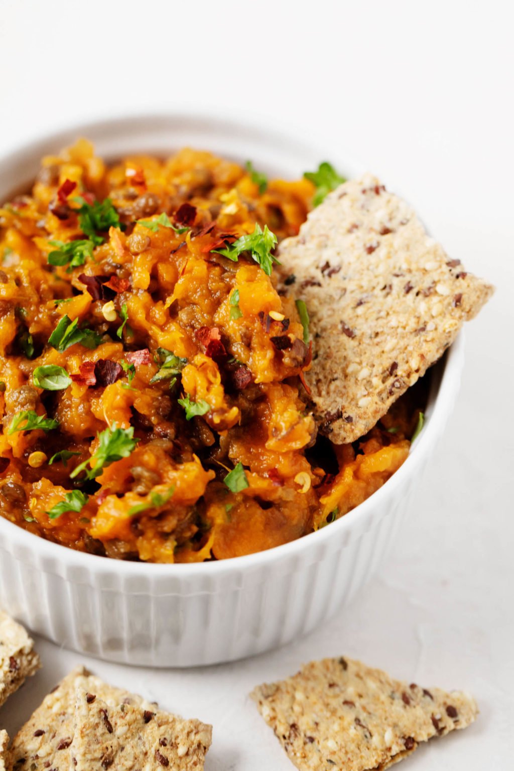 A white ramekin has been filled with a vegan spread made with sweet potatoes and lentils. A single, whole grain cracker is being dunked into the spread.