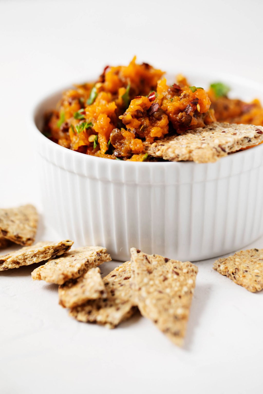 An angled photograph of a white, fluted ramekin filled with an orange dip. Broken crackers lie near the ramekin.