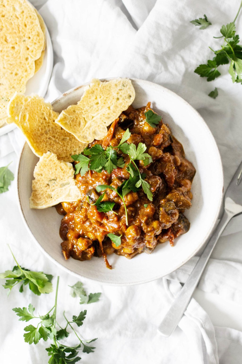 A dish of curried eggplant, tomatoes, and chickpeas is served in a white bowl with a white rim. A silver spoon rests nearby.