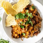 An overhead image of a dish of curried eggplant, tomatoes and chickpeas. Fresh herbs and a flatbread are scattered nearby.