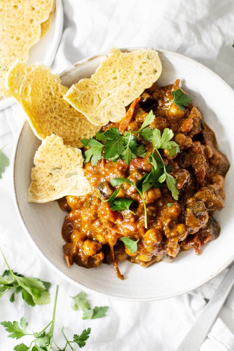 An overhead image of a dish of curried eggplant, tomatoes and chickpeas. Fresh herbs and a flatbread are scattered nearby.