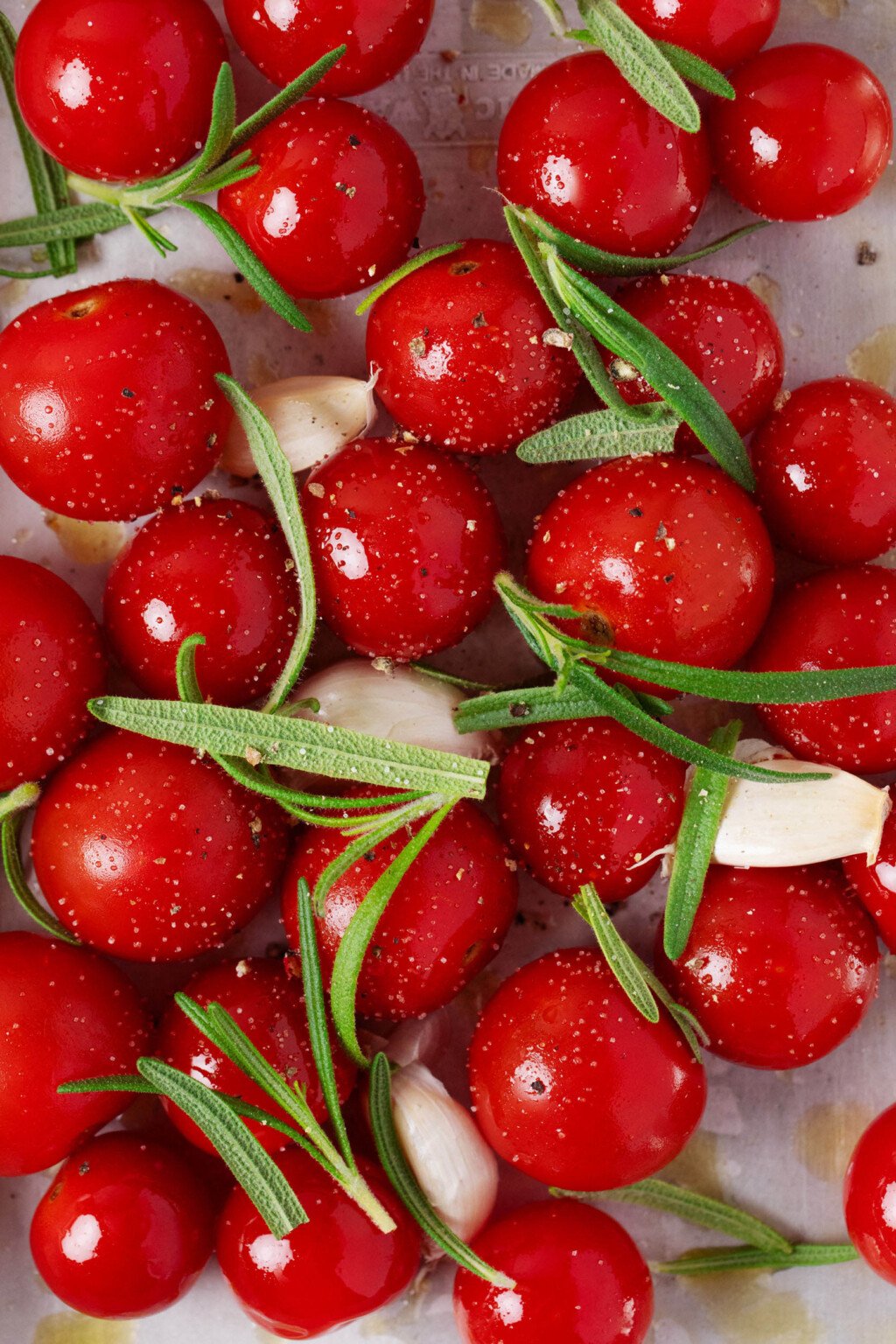 Round, red cherry tomatoes have been mixed with garlic cloves and rosemary leaves.