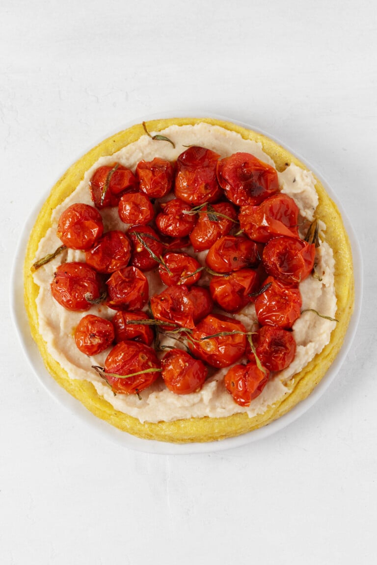 An overhead image of a round polenta tart, which has been topped with oven-roasted cherry tomatoes, herbs, and a spreadable white bean dip.