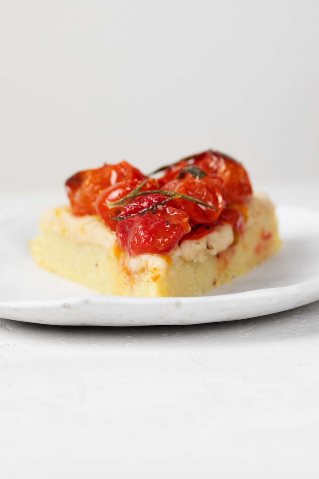 An image of a slice of a polenta tart, which has been topped with oven-roasted cherry tomatoes, herbs, and a spreadable white bean dip.