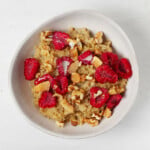 An asymmetrical round bowl is resting on a white surface. It holds a warm breakfast cereal with raspberries, coconut, and walnuts.