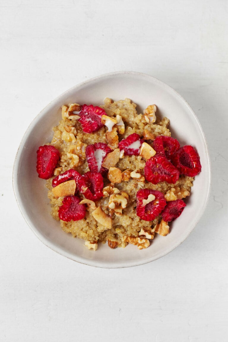 An asymmetrical round bowl is resting on a white surface. It holds a warm breakfast cereal with raspberries, coconut, and walnuts.