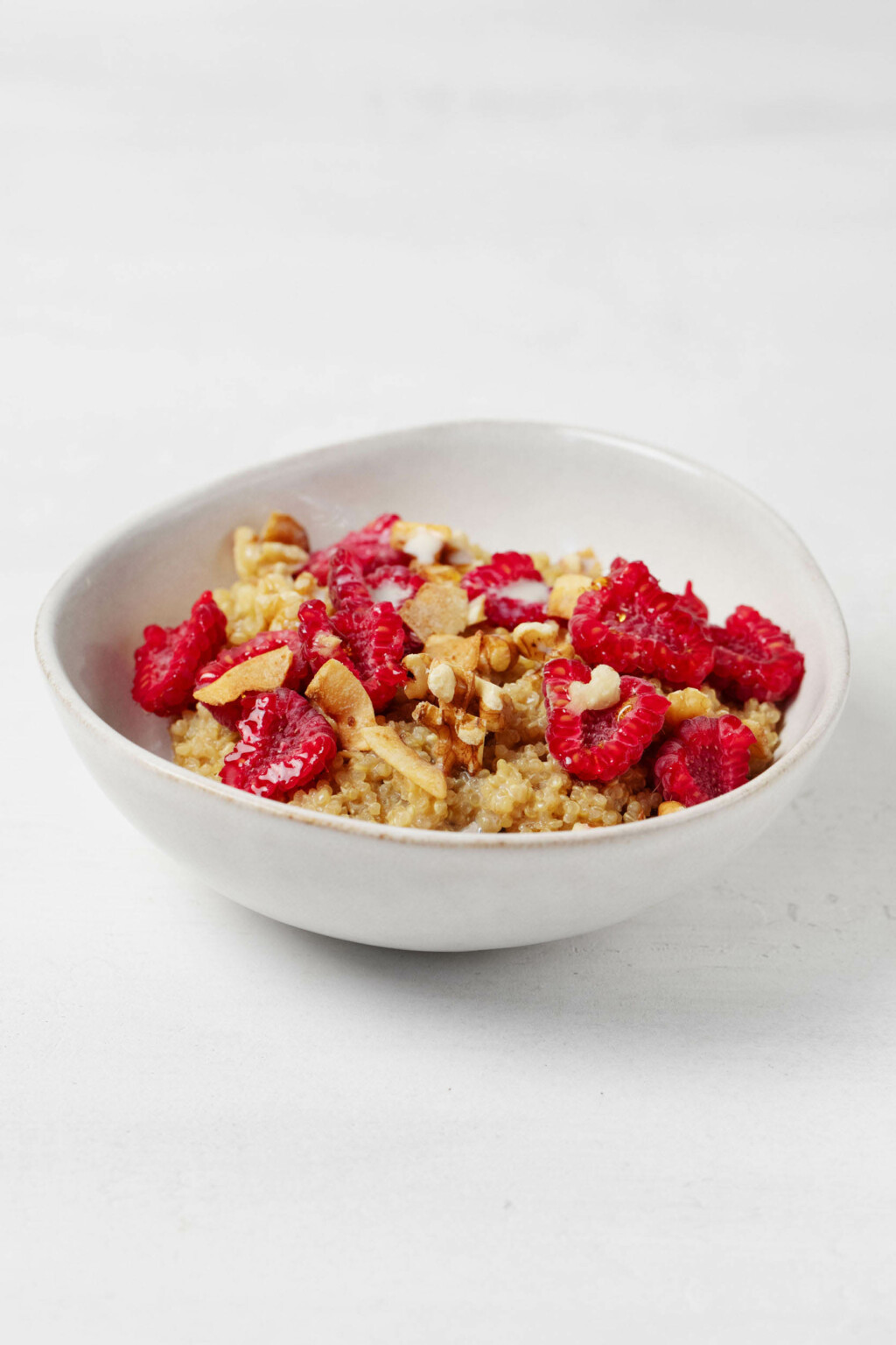 A round bowl is resting on a white surface. It's filled with maple vanilla quinoa porridge, raspberries, and chopped nuts.