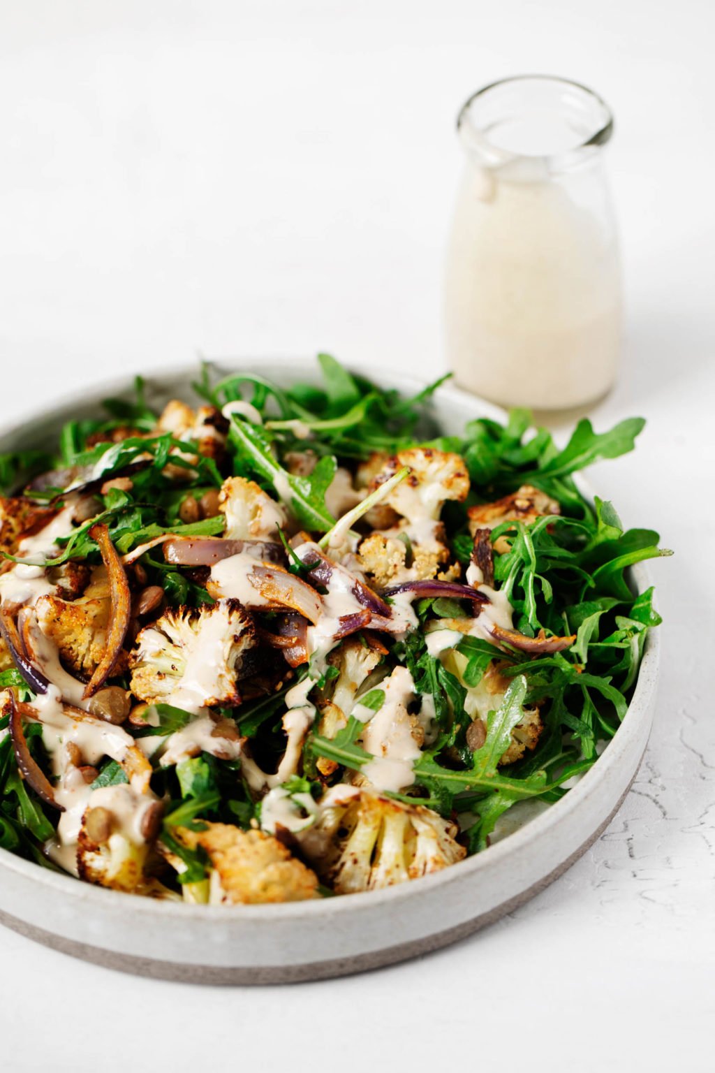 A za'atar roasted cauliflower and green salad has been plated against a white surface. Extra tahini dressing is in a glass container in the background.