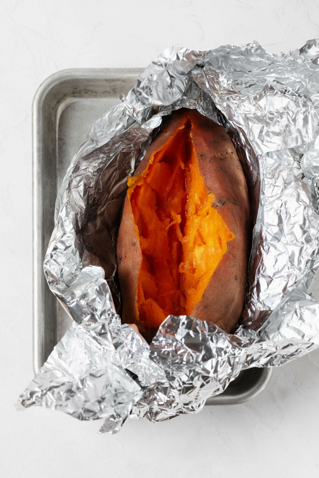 An overhead image of a baked sweet potato, wrapped in foil and resting on a small baking sheet.