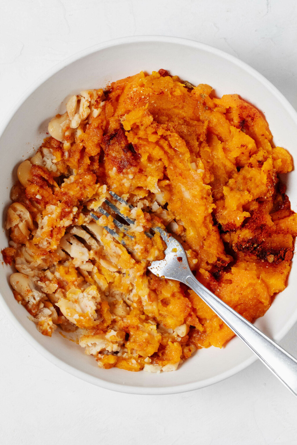 A white round bowl is filled with a mixture of sweet potato and white beans, which is being mashed with a fork.