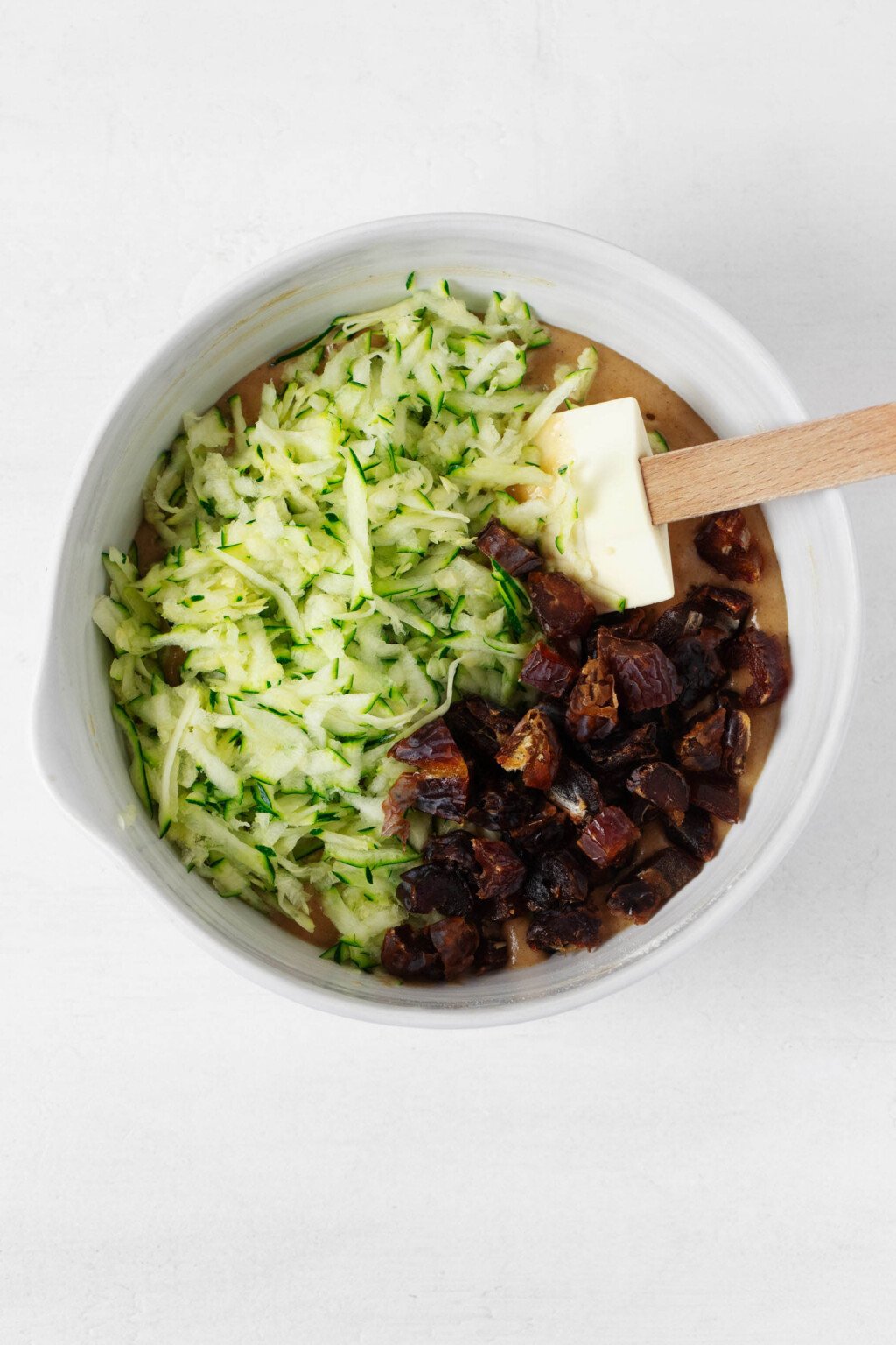 An overhead image of grated zucchini and chopped dates in a round white mixing bowl.