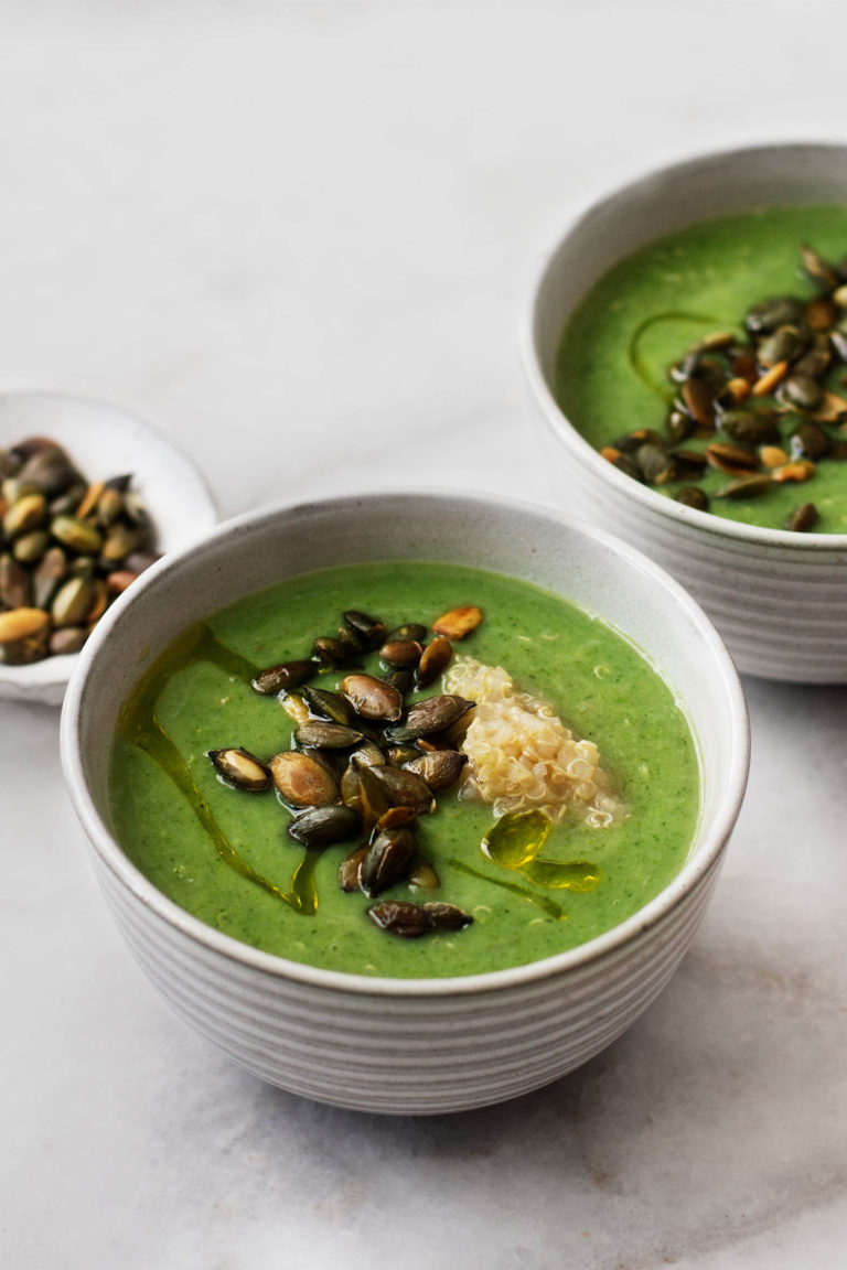 Two bowls of vegan cream of broccoli and quinoa soup, topped with toasted pepitas and a scoop of cooked quinoa.
