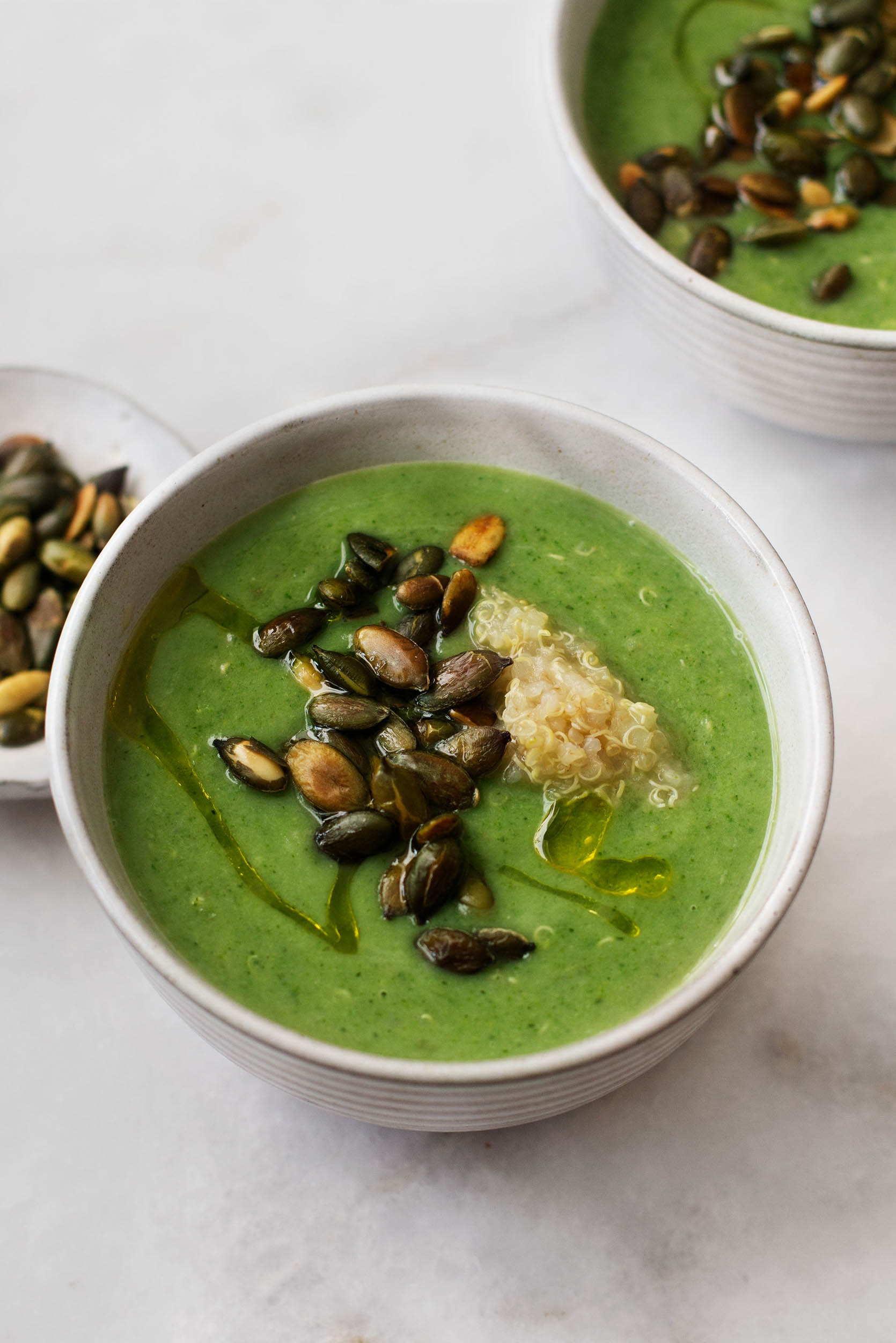 A close up, angled photograph of a bright vegan cream of broccoli and quinoa soup, topped with pepitas.