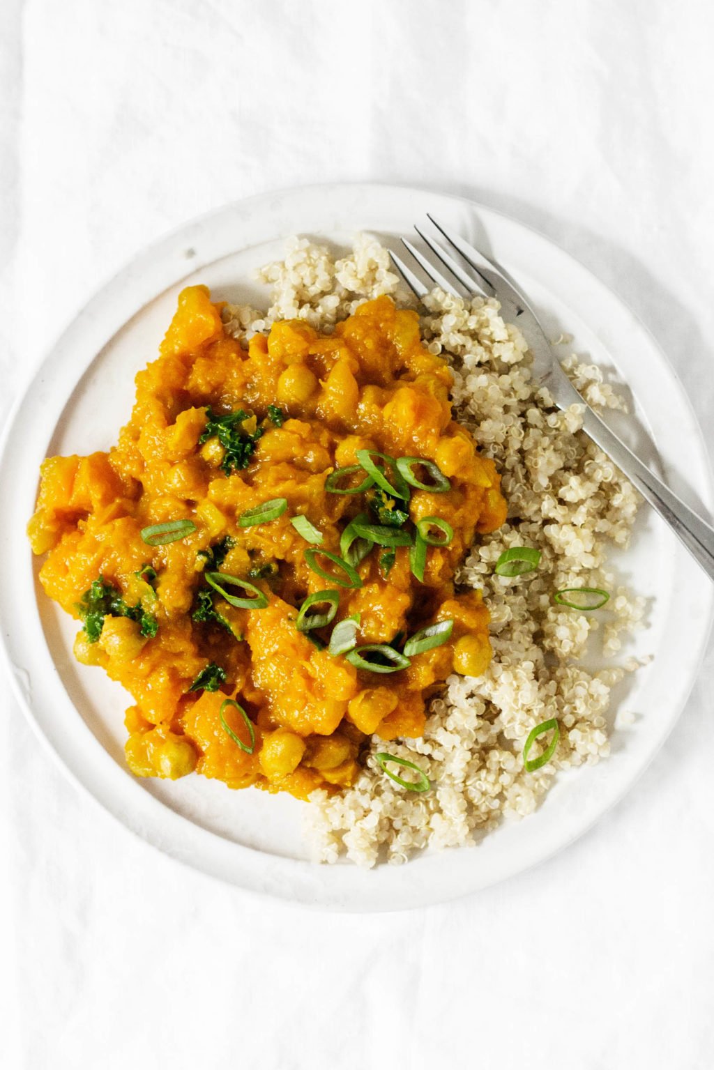 An overhead photograph of a vegan golden butternut squash and chickpea curry, garnished with chopped green onion tops and served with cooked quinoa.