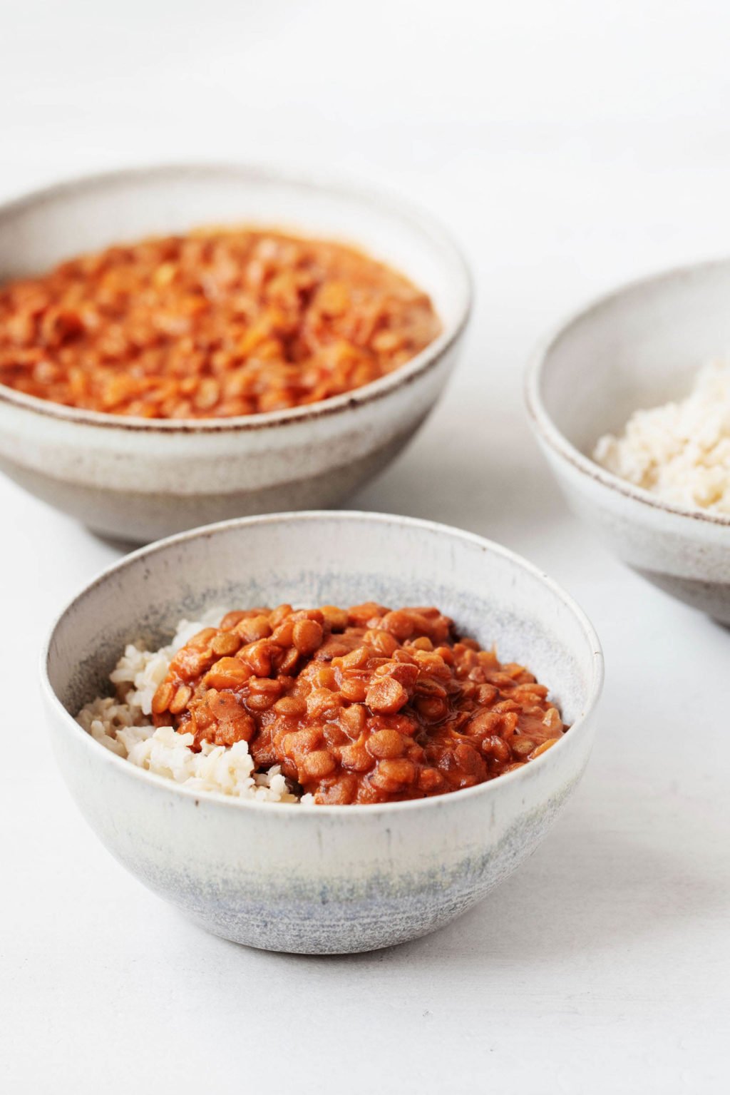 Two ceramic bowls have been piled with masala spiced, cooked lentils. Another bowl in the backdrop is filled with cooked brown rice.