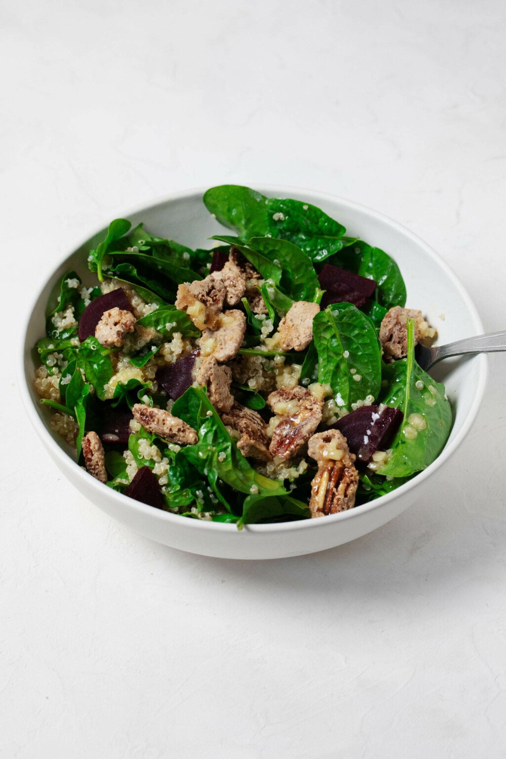 A white ceramic bowl has been filled with roasted beets, baby spinach leaves, quinoa, and candied nuts.
