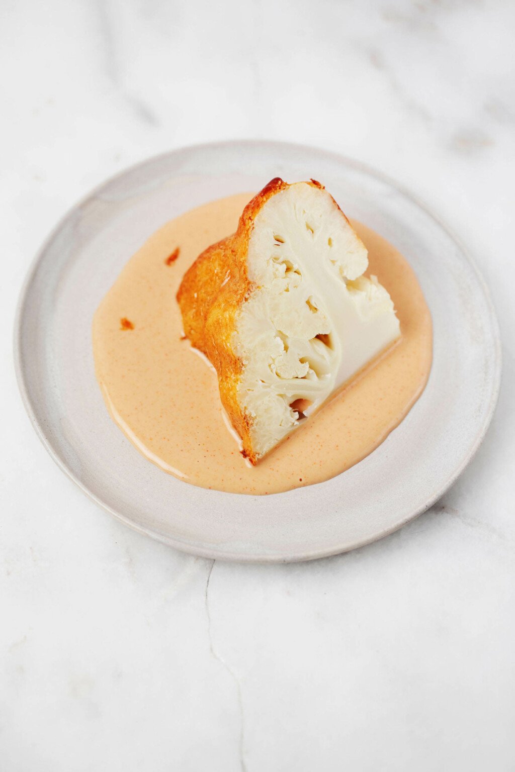 A wedge of whole roasted lemon tahini cauliflower, along with extra sauce, is served on a small, round white plate.