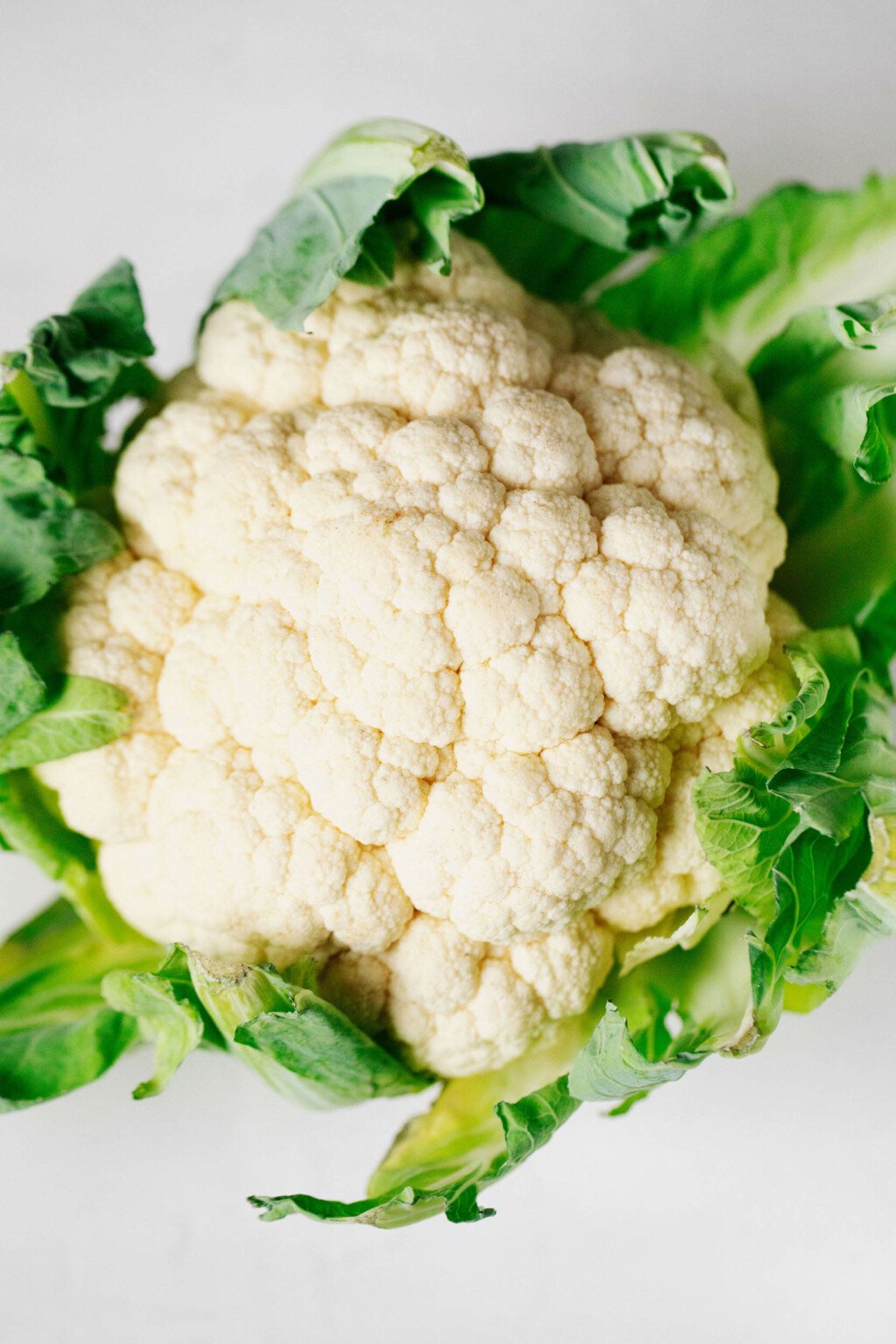 A head of cauliflower, surrounded by green leaves, is resting on a white surface.