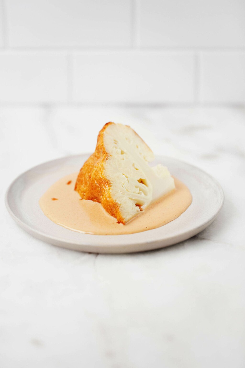 A wedge of whole roasted lemon tahini cauliflower, along with extra sauce, is served on a small, round white plate.