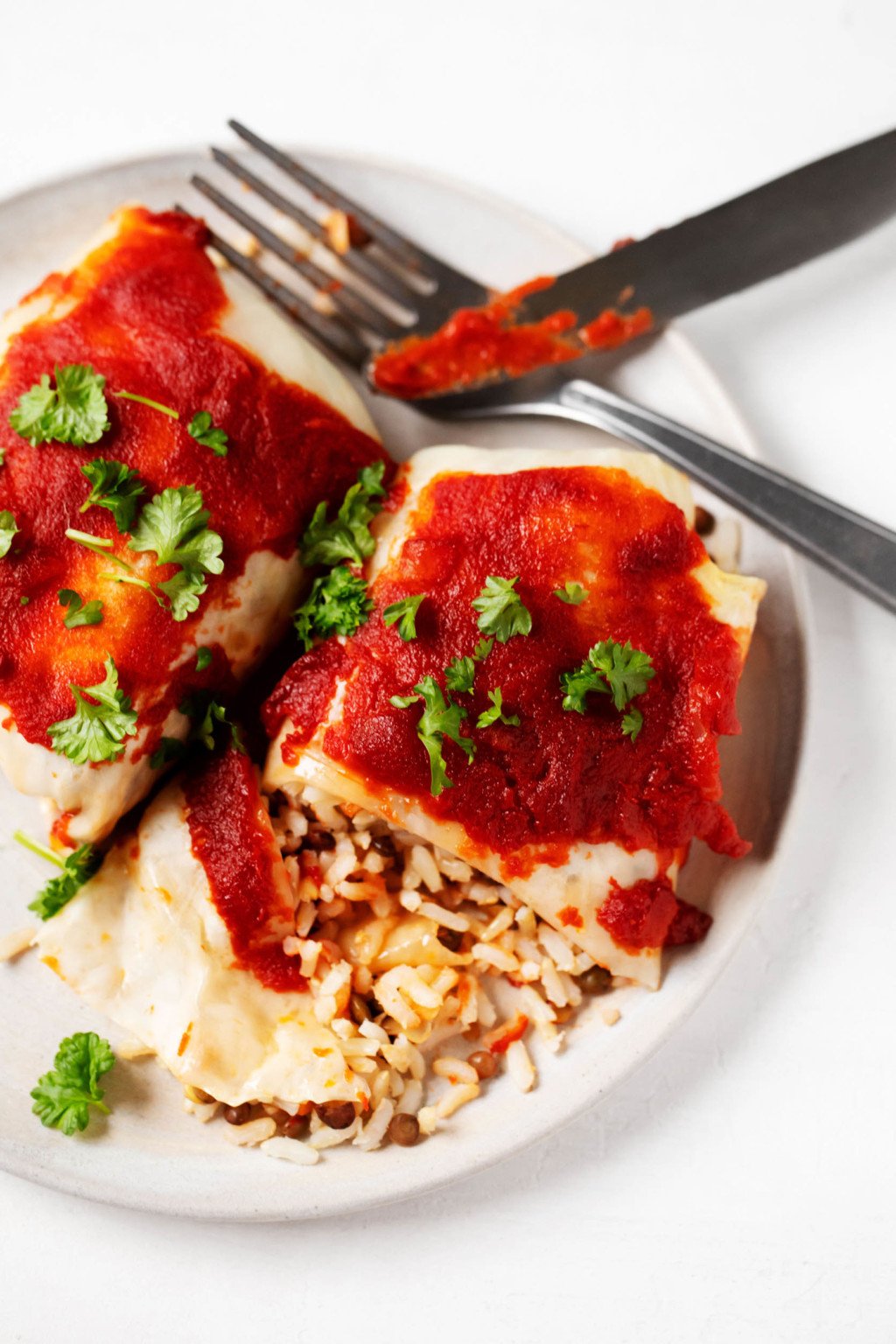 Two stuffed cabbage rolls, covered in tomato sauce, have been laid out on a plate and sliced into with a fork and knife.