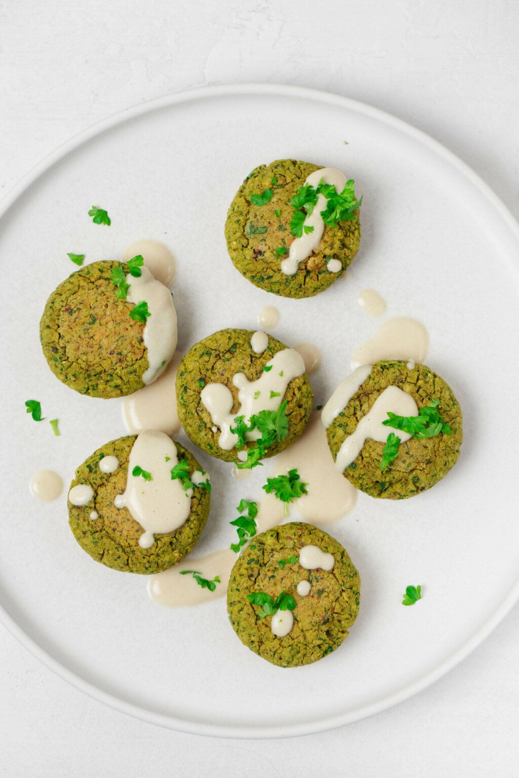 Vegan pistachio spinach falafel have been arranged on a white plate. They're drizzled with tahini sauce and sprinkled with fresh herbs.