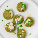 Vegan pistachio spinach falafel have been arranged on a white plate. They're drizzled with tahini sauce and sprinkled with fresh herbs.