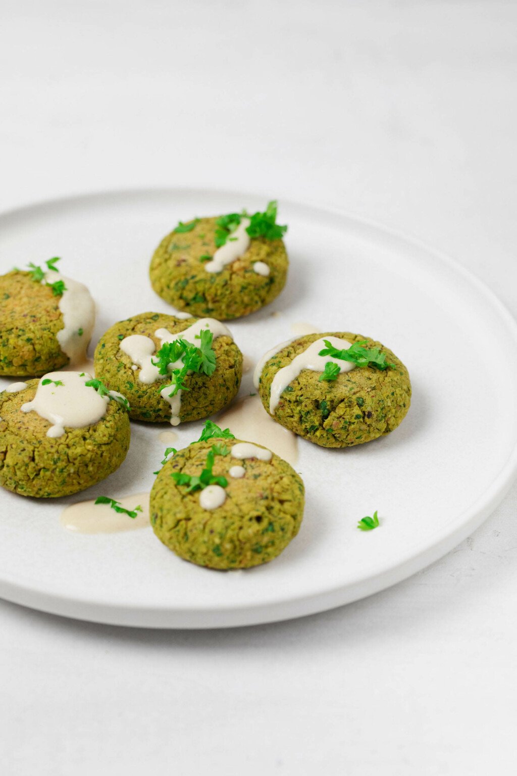 Baked patties of chickpeas, ground pistachios, and spinach have been arranged on a round, rimmed white plate. They're drizzled with a cream colored dressing and sprinkled with fresh herbs.