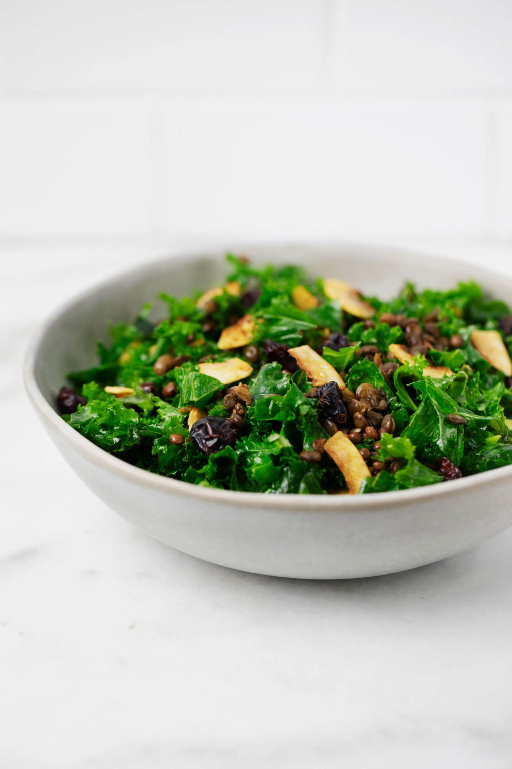 A round, white and gray ceramic bowl holds a festive kale salad with cranberries and coconut bacon.