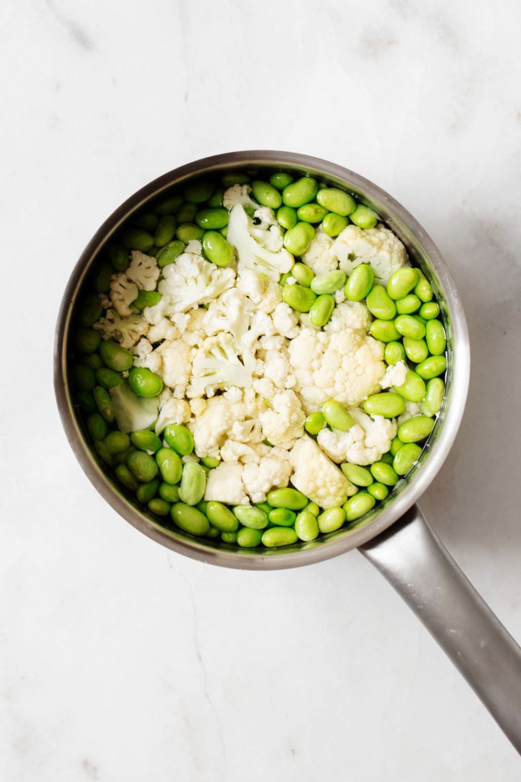 A silver sauce pot is filled with a mixture of shelled edamame and vegetables.