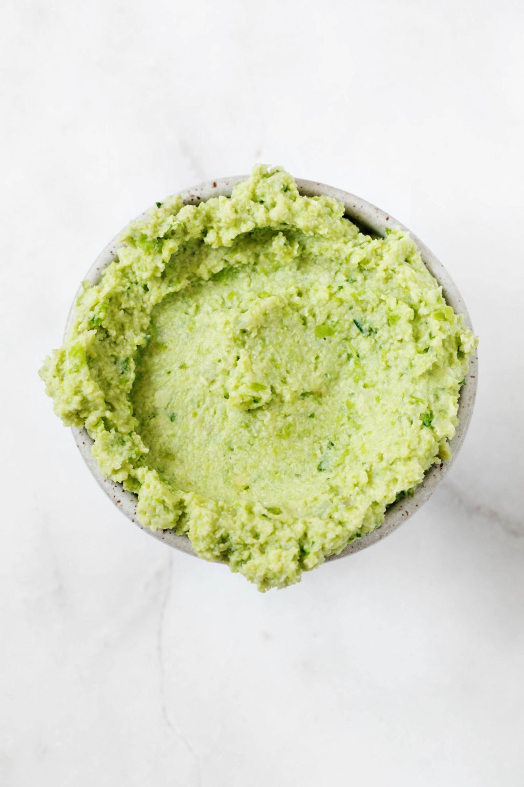 A small, white pinch bowl of pureed edamame rests on a white marble surface.