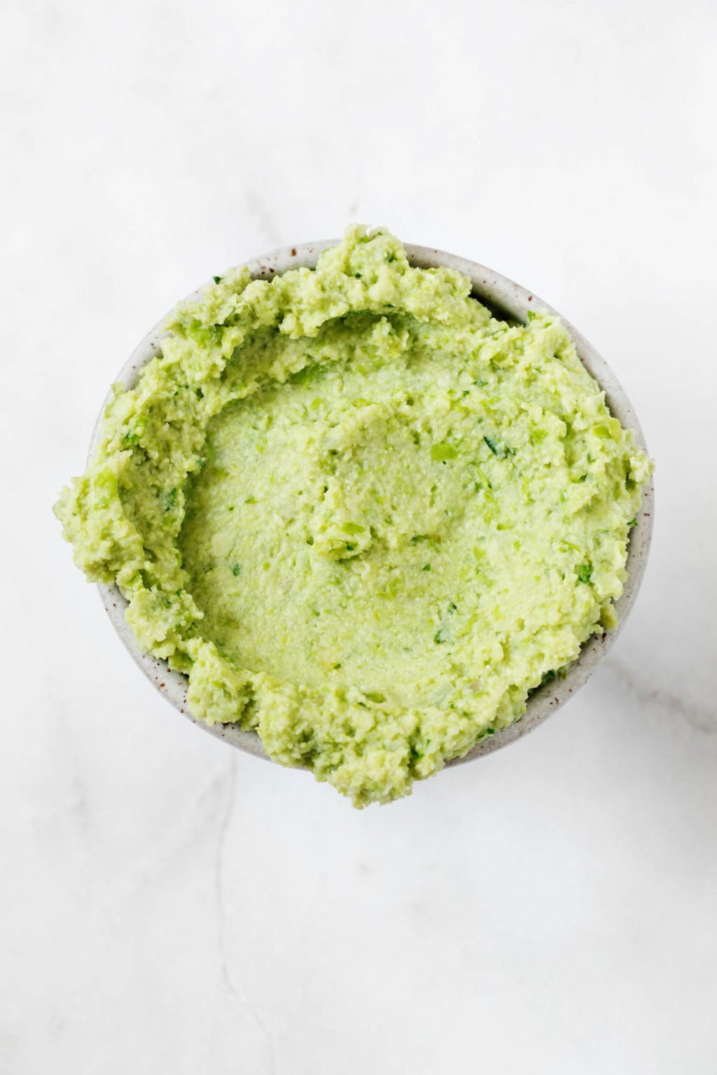 A small, white pinch bowl of pureed edamame rests on a white marble surface.