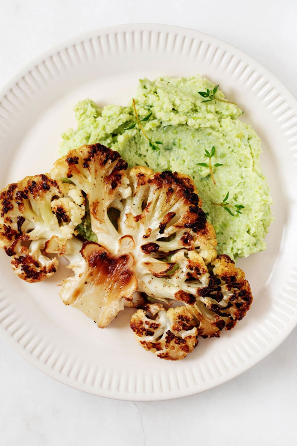 A seared cauliflower steak has been served with a light green edamame mash on a rimmed, cream colored serving plate.