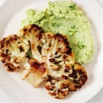 A seared cauliflower steak has been served with a light green edamame mash on a rimmed, cream colored serving plate.