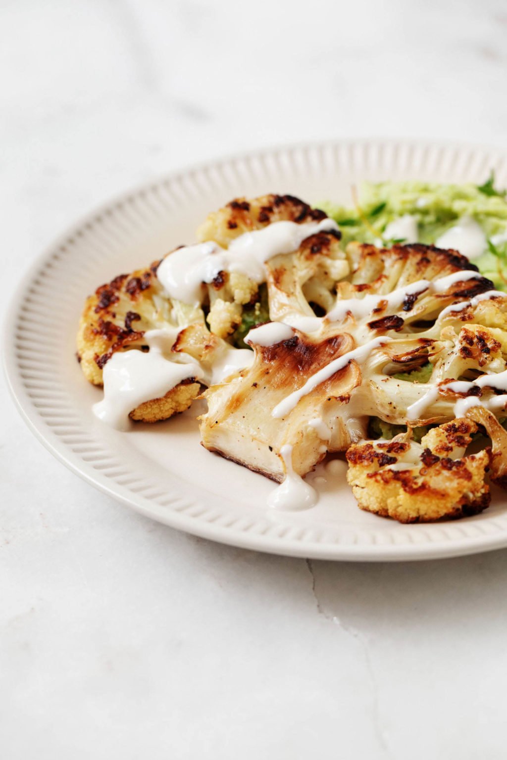 An angled photograph of a white, rimmed plate with a seared slab of cauliflower and a small accompanying portion of mashed green edamame.